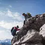 Zwei Wandernde klettern an einem felsigen Berghang: Eine Person in roter Jacke steigt von unten hinauf, während eine andere mit grünem Rucksack oben steht und hilft. Im Hintergrund sind ein weiter Himmel mit Wolken sowie schneebedeckte Berggipfel zu sehen.