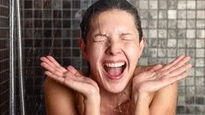 Young woman reacting in shock to hot or cold shower water as she stands under the shower head washing her hair eyes closed with her hands raised and mouth open