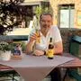Ein Mann sitzt an einem Tisch, lächelt in die Kamera und hält ein Glas Bier in der rechten Hand. Auf dem Tisch steht eine Flasche Bier.