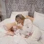 Eine Frau und ein Baby, beide tragen weiß, liegen in einem Bett. Die Frau kämmt das Baby