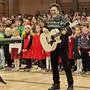 Mann mit heller Western-Gitarre, dahinter zahlreiche Volksschulkinder | Peter Karpf mit seinem Kinderchor