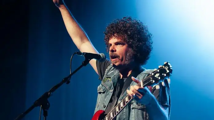 Der Sänger ist in Jeans gekleidet und hat eine Wuschelfrisur. Er hat eine rote Gitarre umgehängt und reckt den rechten Arm in die Luft