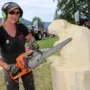 Eine Frau mit einer Motorsäge arbeitet an einem Holzblock. Sie lächelt in die Kamera.