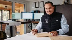 Ein Mann mit dunklen Haaren trägt ein helles Hemd mit einer sportlichen Weste darüber, in der Hand hat er ein Stück Holz und lächelt in die Kamera