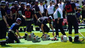 Mehrere Personen mit Feuerwehruniform und Helm absolvieren einen Wettkampf. Vor ihnen liegen eingerollte Wasserschläuche.