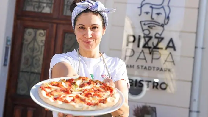 Eine Frau hält einen weißen Teller mit einer Pizza in die Kamera und lächelt