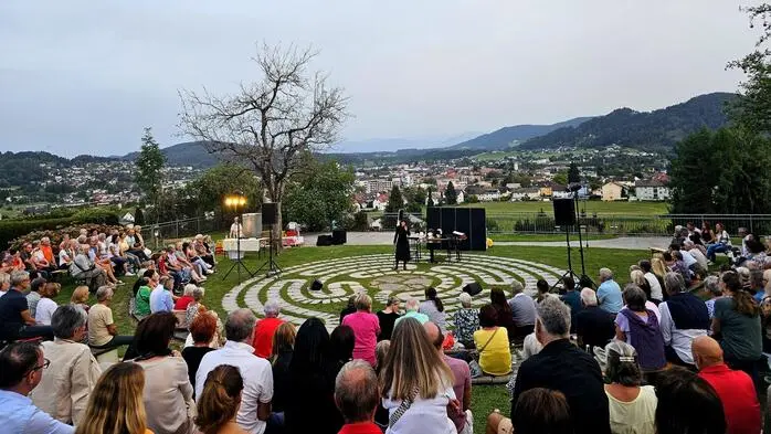 Ein Open-Air-Kabarett hoch über Feldkirchen. Zahlreiche Menschen sitzen um eine Bühne.