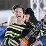 Harry Styles steht auf einer Bühne und singt mit offenem Mund, während er eine Gitarre spielt. Er trägt einen gestreiften Pullover, im Hintergrund sind weitere Musiker und Bühnenlichter zu sehen. | Die neue Single von Harry Styles startet mit tanzbaren Beats. 