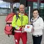 Ein Mann in Rettungsuniform bekommt einen Defibrillator von einer Frau in weißer Jacke und Seidenschal überreicht