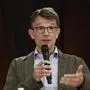 Pointner gestikuliert mit einer Hand, die andere hält ein Mikrofon. Er trägt einen braunen Blazer mit hellem Hemd darunter, eine Brille im Gesicht