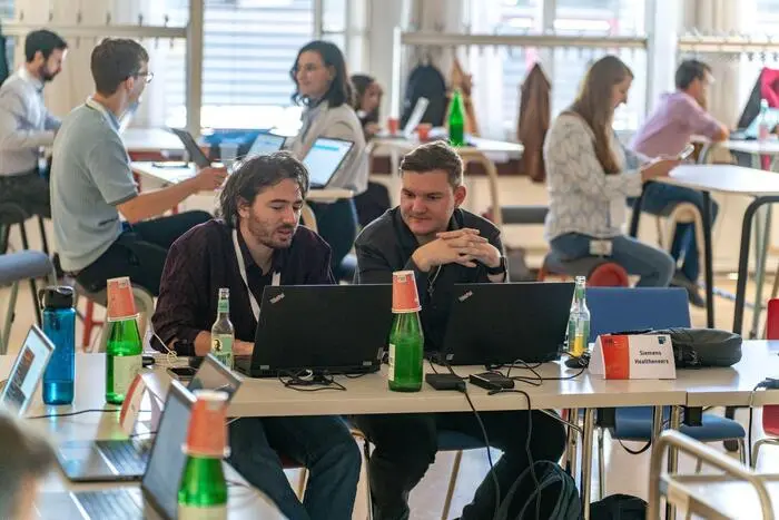 Zwei männliche Personen sitzen vor ihren aufgeklappten Laptops und sprechen miteinander.