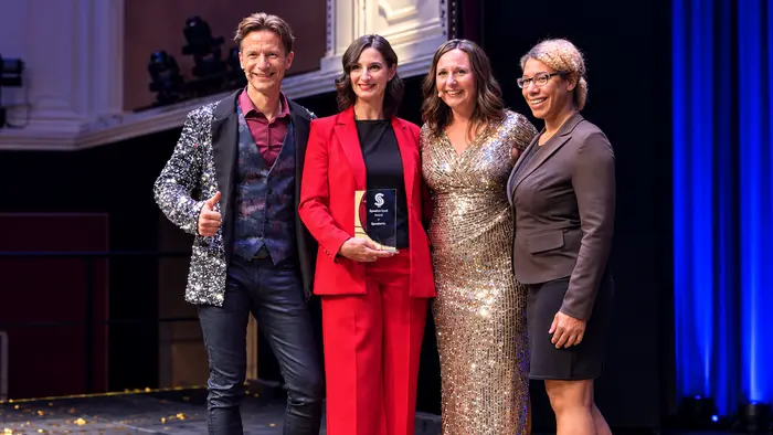 Drei Frauen und ein Mann stehen in eleganter Abendkleidung auf einer Bühne und stehen für ein Foto bereit.