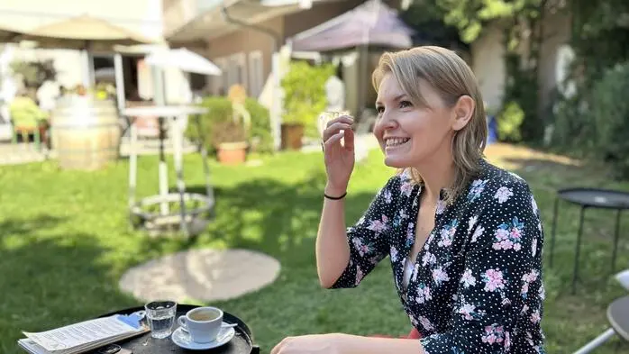 Eine Frau in sommerlicher Kleidung sitzt in einem Garten und spricht lächelnd
