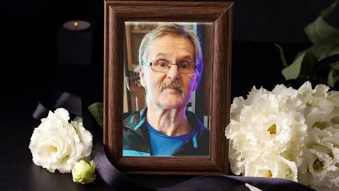 Ein Bild von einem Mann mit Brille steckt in einem braunen Bilderrahmen, daneben liegen weiße Blumen