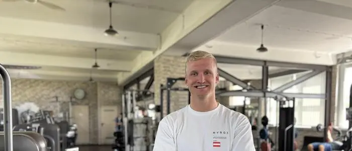 Ein blonder Mann in weißem Shirt steht in einem Fitnessstudio und lächelt in die Kamera