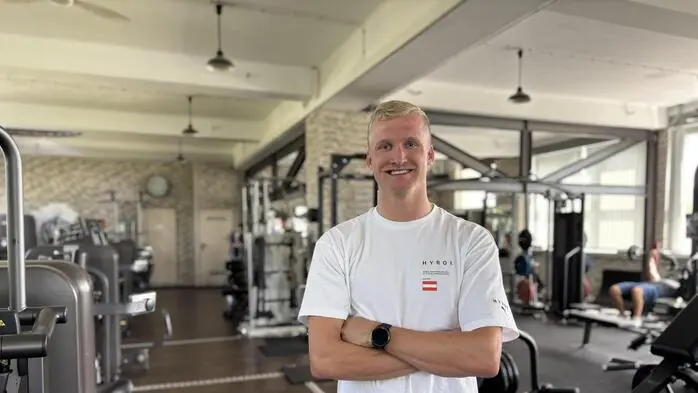 Ein blonder Mann in weißem Shirt steht in einem Fitnessstudio und lächelt in die Kamera