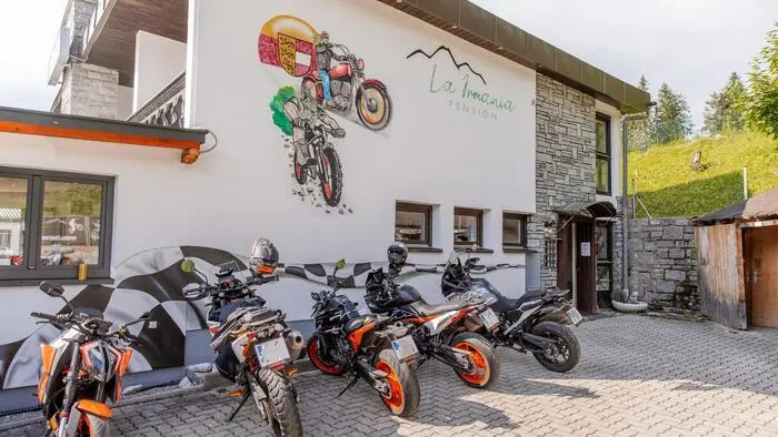 Mehrere Motorräder stehen vor einem hotelähnlichen Gebäude, auf dem zwei Motorräder gezeichnet sind und das Logo der Pension ersichtlich ist