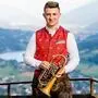 Ein junger Mann in Tracht hälte eine Trompete in der Han dund lächelt in die Kamera, im Hintergrund Berge und ein See