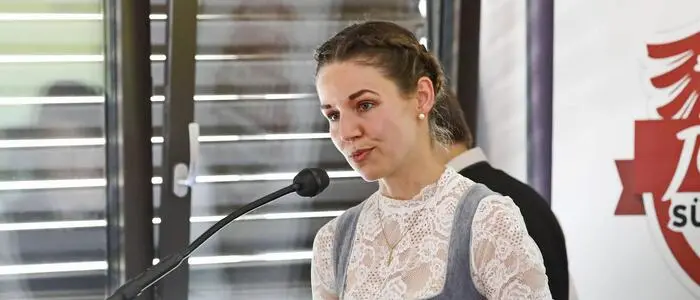 Melanie Mair spricht in ein Mikrofon. Hinter ihr ist ein gekipptes Fenster mit heruntergelassener Jalousie.
