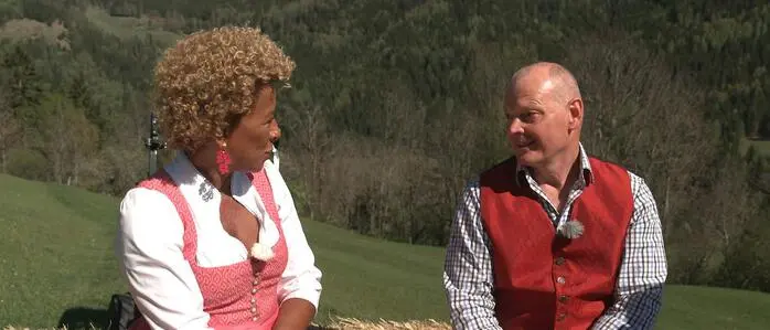 Ein Mann und eine Frau, beide in Tracht, sitzen auf einem Strohballen und unterhalten sich 