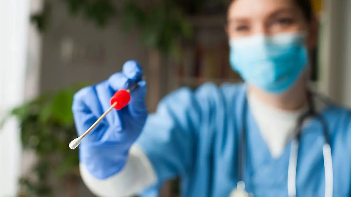 Female caucasian NHS doctor holding a swab collection stick, nasal and oral specimen swabbing in doctor's office, patient PCR testing procedure appointment, Coronavirus COVID-19 global pandemic crisis