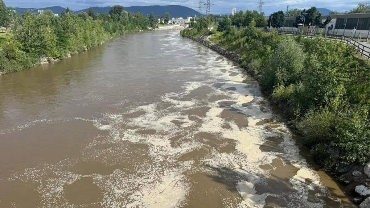 Blick auf das braune Wasser der Mur in Richtung Norden, an der Oberfläche schwimmen weiße Schaumflächen, im Hintergrund ist das Grazer Murkraftwerk zu sehen