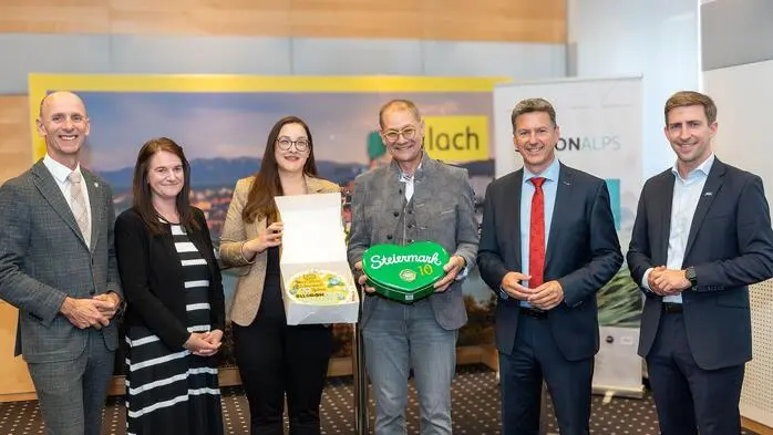 Mehrere Personen stehen bei einer Pressekonferenz im Halbkreis und halten eine Torte in die Kamera