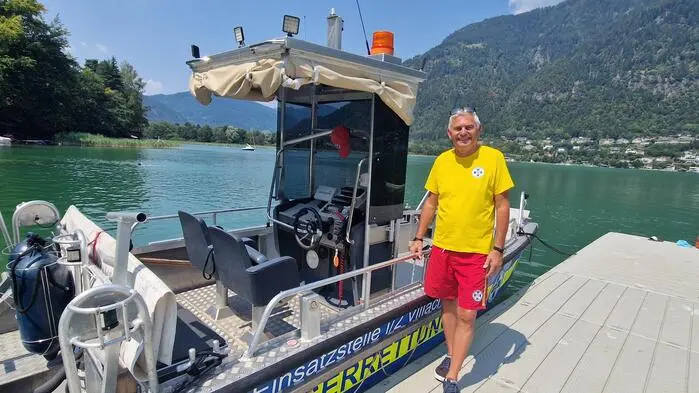 Ein Mann in roter Badehose und gelbem T-Shirt steht auf einem Steg neben einem Einsatzboot der Wasserrettung