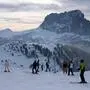 Das Bild zeigt eine winterliche Berglandschaft mit einer Gruppe von Skifahrern, die sich auf einer Skipiste befinden. Im Hintergrund erstrecken sich verschneite Berge, darunter ein markanter, felsiger Gipfel. Die Landschaft ist von schneebedeckten Hügeln und verstreuten Nadelbäumen geprägt. Der Himmel ist teilweise bewölkt, was eine leicht gedämpfte Beleuchtung erzeugt. Einige Skifahrer stehen still, während andere sich auf die Abfahrt vorbereiten. Links im Bild ist eine Seilbahn mit einer Gondel zu sehen. Die Aufnahme zeigt die Sellaronda im Skigebiet Dantercepies mit Blick auf den Langkofel. | Die Sellaronda im Skigebiet Dantercepies mit Blick auf den Langkofel.