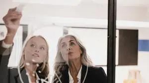 Mature and young businesswoman drawing chart on glass pane in office
