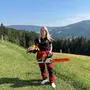 Eine blonde Frau steht in einer Schnittschutzhose und einer Motorsäge in der Hand auf einem Berg