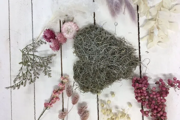 Wenn das Herz fertig umwickelt wurde werden die Trockenblumen gekürzt. und auf eine Länge von je ca. 5 bis 10 cm gebracht. Nun das Herz mit den ausgesuchten Trockenblumen dekorieren. Dafür einfach die Stielenden in das Moos stecken oder mit einer Heißklebepistolen festkleben. Am Ende kann zusätzlich noch ein Draht eingefädelt werden, an dem man das Herz später aufhängen kann.