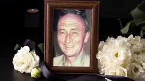 Ein Foto von einem Mann in einem dunkelbraunen Rahmen umgeben von weißen Blumen und einer schwarzen Schleife