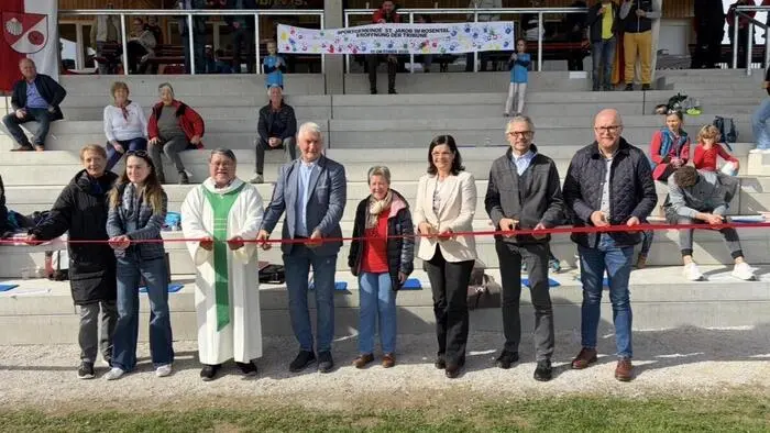 Mehrere Personen stehen vor einer Zuschauertribüne eines Sportplatzes und schneiden ein rotes Band durch