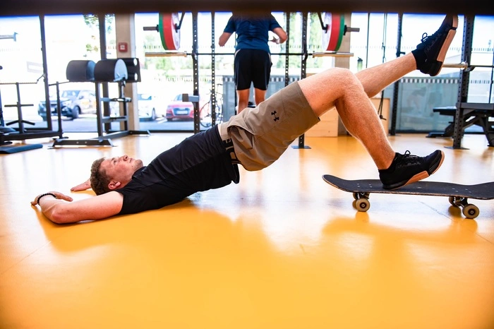 Kräftigung der hinteren Muskelkette (unterer Rücken, Hüfte, Oberschenkel Rückseite) mit Hilfe eines Skateboards: Hüftbrücke mit beiden Beinen auf dem Skateboard, ein Bein wird gehoben und mit dem anderen Bein bei voller Hüftspannung das Skateboard langsam nach vorne geführt.