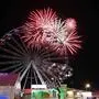 Eine Feuerwerksrakete explodiert am Nachthimmel, davor sieht man das Riesenrad eines Vergnügungsparks