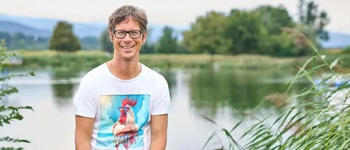 Mann mit braunen Haaren und Brille sitzt auf einem Geländer, lacht in die Kamera und hinter ihm ist ein See