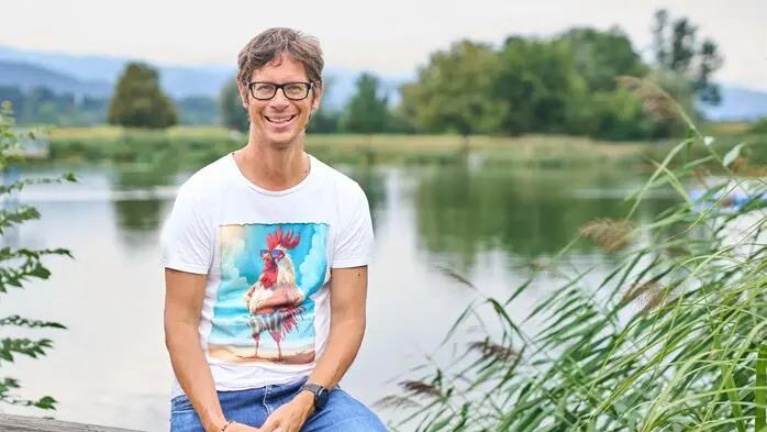 Mann mit braunen Haaren und Brille sitzt auf einem Geländer, lacht in die Kamera und hinter ihm ist ein See