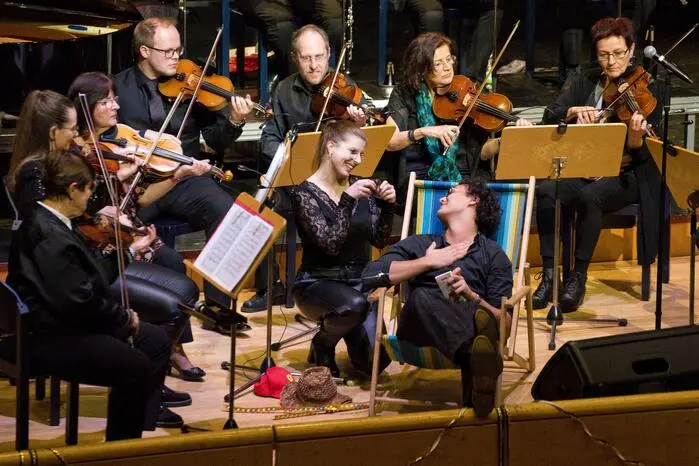 Als Konzertmeisterin führte Lore Schrettner persönlich durch den Abend und kommentierte die einzelnen Werke. Mit der „Schlaraffen-Polka“ sorgte das Orchester für ein heiteres Bühnenbild: Kulinarische Genüsse schwebten von der Decke herab, während ein Protagonist im Liegestuhl verwöhnt wurde. 
