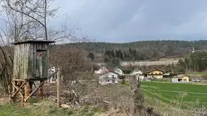 Blick auf eine Häusergruppe hinter einem Feld. Im Vordergrund steht ein Hochstand, links davon kleinere Bäume