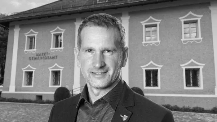 Portät eines Mannes in schwarz-weiß vor einem zweistöckigen Gebäude mit der Aufschrift „Marktgemeinde Treffen“