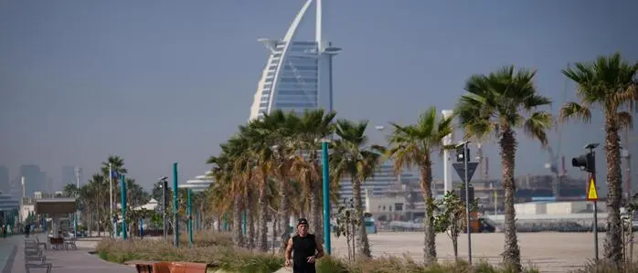 Das Bild zeigt das Luxushotel Burj Al Arab in Dubai, Palmen und eine Promenade mit einem Läufer im Vordergrund. 