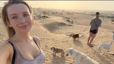 Eine blonde Frau steht mit ihren Hunden in der Wüste und lächelt in die Kamera