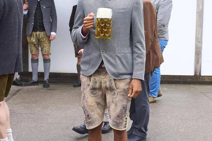 Michael Olise mit Trachtensakko und Lederhose. Die Schuhbänder lässig offen...