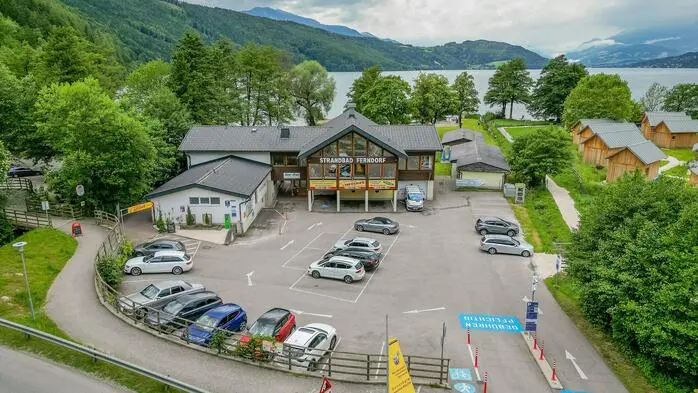 Drohnenaufnahme eines Parkplatzes der gut besucht ist, im Hintergrund sieht man Berge und einen See