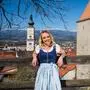 Eine junge Frau mit blonden Haaren lehnt sich an einen Holzzaun, im Hintergrund ist der Kirchenturm von Wolfsberg zu sehen 