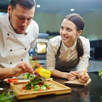 Young woman writing down recipe of vegetable salad while chief cooking it