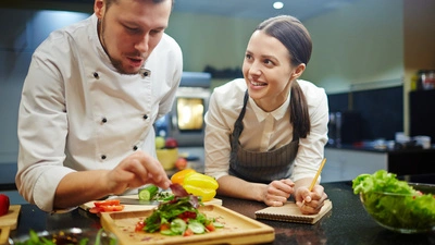 Young woman writing down recipe of vegetable salad while chief cooking it