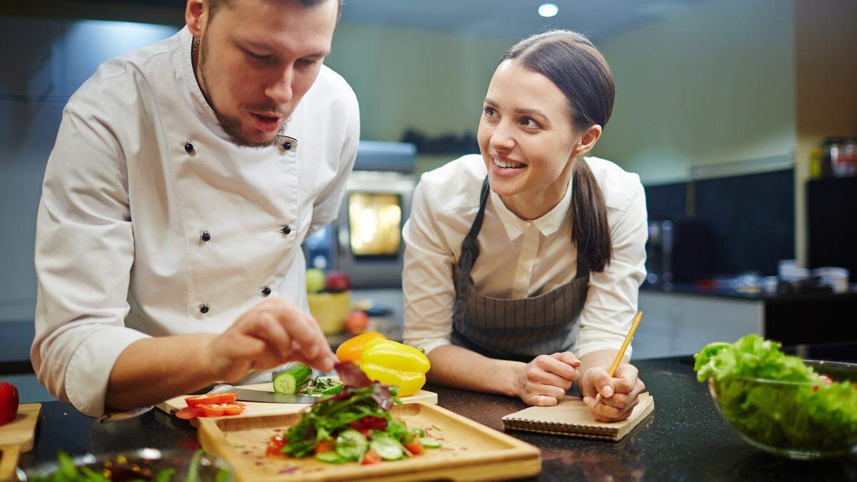 Young woman writing down recipe of vegetable salad while chief cooking it