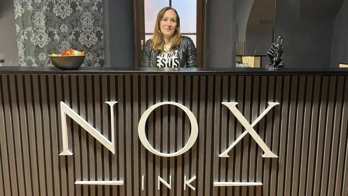 Eine Frau steht hinter einer Theke. Die Wand im Hintergrund ist schwarz. Auf der Theke steht Nox Ink.